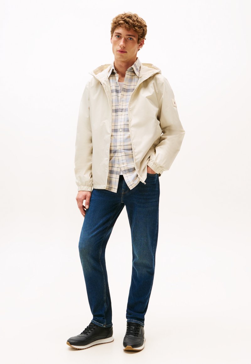 Giacca beige chiaro con cappuccio e polsini elasticizzati, indossata sopra una camicia a scacchi blu e bianca. Jeans blu scuro e sneakers nere completano l'outfit.