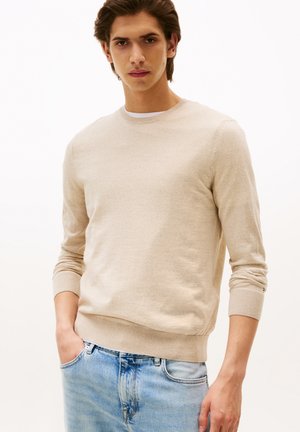 Maglione beige a maglia con scollo rotondo e maniche lunghe, dotato di polsini e orlo a costine. Indossato con jeans azzurri chiari.
