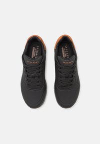 Adidași negri pentru sport, cu detalii perforate, din material sintetic texturat, pun accent pe un toc maro și logo-ul Skechers pe limbă.