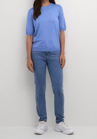 Donna che indossa un maglione azzurro a maniche corte, jeans blu e sneakers bianche, in piedi davanti a uno sfondo grigio chiaro uniforme.