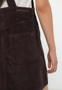 Brun cordfløyelsoverall med justerbare stropper. Har en frontlomme med broderte detaljer og et lite kontrasterende logokort.
