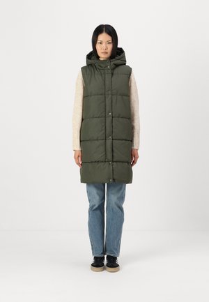 Kaki puffervest med hette, med knappelukking og quiltet mønster, kombinert med lyseblå jeans og svarte sneakers.