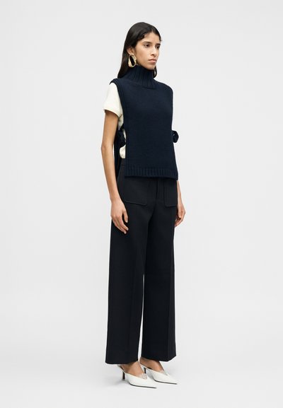 Μπλε ναυτικό πλεκτό γιλέκο με ψηλό λαιμό φορεμένο πάνω από λευκό t-shirt, σε συνδυασμό με μαύρο, ψηλόμεσο παντελόνι φαρδιάς γραμμής και λευκές ψηλοτάκουνες μυτερές γόβες.