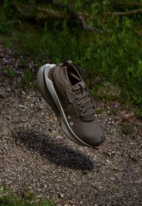 Chaussure de sport marron avec une tige texturée, une semelle superposée et des accents réfléchissants, flottant au-dessus d'un chemin de gravier avec de la verdure en arrière-plan.