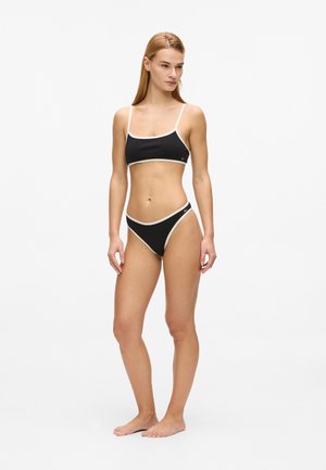 Giovane donna in piedi a piedi nudi che indossa un bikini nero con bordi bianchi su uno sfondo bianco semplice.