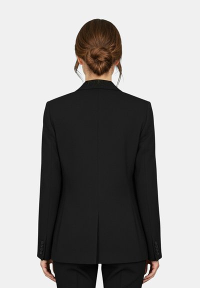 Donna con capelli castani raccolti in uno chignon, indossa un blazer nero su misura e pantaloni neri abbinati, in piedi di spalle contro uno sfondo neutro.