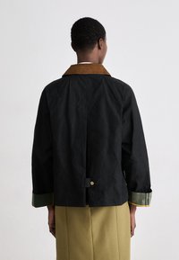 Casaco preto cropped com colarinho de veludo cotelê castanho, detalhes nas mangas em verde e amarelo, e um botão na parte de trás. Tecido texturizado.