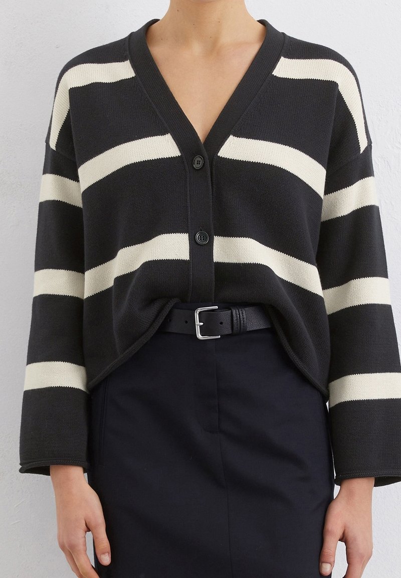 Schwarz- und cremegestreifter Cardigan mit V-Ausschnitt, vier Knöpfen und lockeren Ärmeln, getragen mit einem schwarzen Gürtel und einem Rock mit hoher Taille.