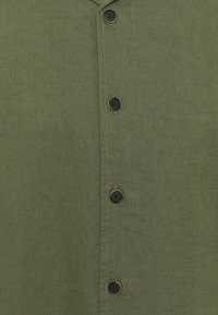 Camicia in lino verde oliva con chiusura a bottoni frontale composta da quattro bottoni neri, uno dei quali ha un accento giallo, e un colletto strutturato.