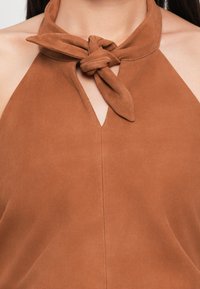 Brun halter-top laget av myk semsket skinn med en knyttet nakkebånd og en sentral nøkkelhulldetalj, med en glatt tekstur og en tettsittende silhuett.