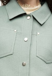 Gros plan d'une veste en laine vert clair avec des boutons-pression argentés et deux poches avant, portée sur un col de haut crème orné de dentelle.