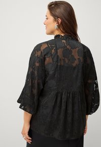 Sort bluse med blomstret blondeoverlay, gennemsigtige lange ærmer, volang ved halsen og peplumskæring. Tekstureret med indviklede detaljer.