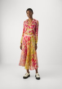 Blusa traslucida con motivo floreale e colletto arricciato, abbinata a una gonna a pieghe dai colori vivaci, rosa, giallo e verde. Scarpe bianche spesse.