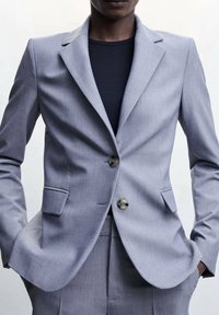 Blazer grigio chiaro su misura con rever stretto, due tasche anteriori, chiusura con un solo bottone e texture del tessuto liscia.