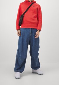 Röd huvtröja med en svart logotyp, lösa blå denimjeans och vita sportskor. Inkluderar en liten svart axelväska.