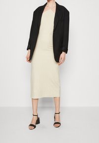 Béžové pletené šaty s vertikálním žebrováním, nošené pod černým oversized blejzrem. Černé sandály na vysokém podpatku celý outfit doplňují.