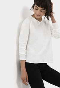 Witte lange mouwen shirt met een relaxte pasvorm en textuurstof. Heeft raglanmouwen en een brede halslijn. Gepaard met zwarte jeans.