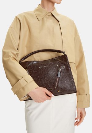 Osoba v béžové bundě a bílých kalhotách drží tmavě hnědou koženou kabelku se zipem a logem „BADURA“.