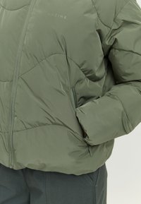 Olive green puffer jacket with quilted texture, zippered side pocket, and MAZINE logo. Features a stand-up collar and lightweight material.