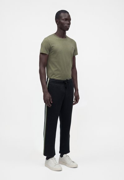 Homme debout portant un t-shirt vert olive, un pantalon de survêtement noir avec des bandes latérales et des baskets blanches sur un fond blanc uni.