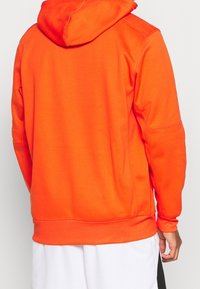 Sweat à capuche orange avec un cordon de serrage à la capuche, des poignets côtelés, et une texture unie. Le dos présente un design sans coutures, sans logos ni motifs.