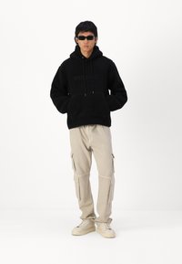 Felpa nera in pile con tasca a marsupio e lettering in rilievo, abbinata a pantaloni cargo beige con più tasche e sneakers chiare.