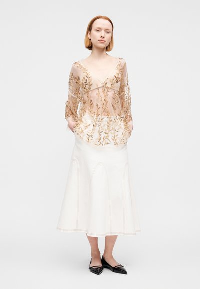 Διάφανο, γυμνό τοπ με χρυσή κεντητική λουλουδιών· λευκή φούστα με φαρδιά γραμμή και καμπυλωτές ραφές· μαύρα μυτερά φλατ παπούτσια· μινιμαλιστικό φόντο.