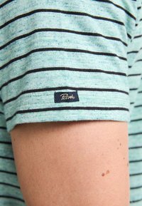 Lichtblauw shirt met korte mouwen en zwarte horizontale strepen. Heeft een klein zwart label met "Petzel" op de mouw. Zachte textuur.