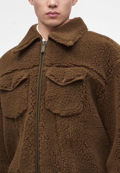 Personne portant une veste polaire marron texturée avec une fermeture éclair frontale et deux poches poitrine à rabats sur un fond uni.