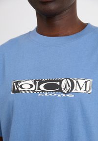 T-shirt din bumbac albastru, cu un grafic central având cuvintele "VOLCOM stone" în alb și negru. Guler rotund și mâneci scurte.