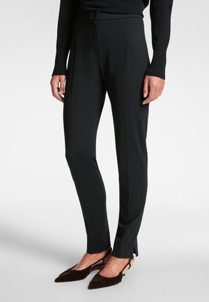 Pantalones negros ajustados con una tela suave, que presentan un cierre de botón y aberturas en los laterales del dobladillo. Llevados con zapatos de terciopelo marrón.