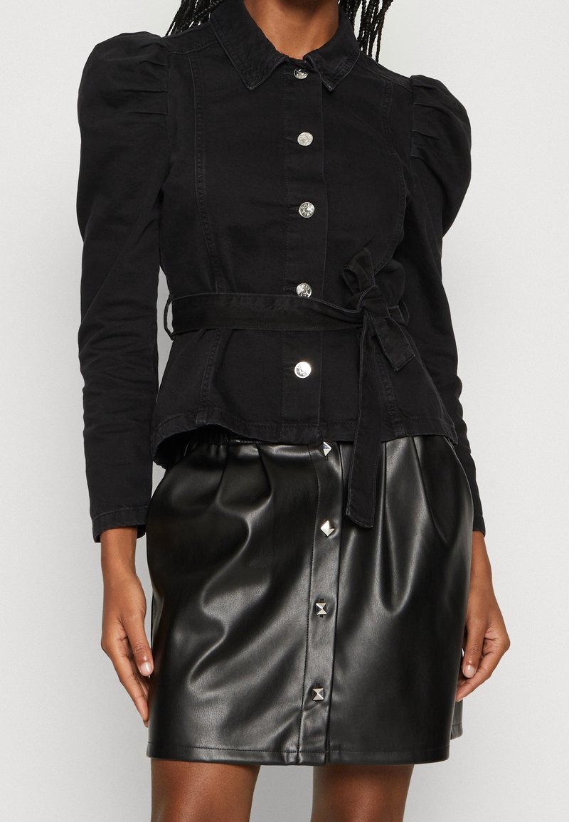 Nainen, jolla on yllään musta nappijakku, jossa on puffihihat ja sidottu vyö, yhdistettynä mustiin nahkahousuihin, joissa on hopeanvärisiä nappeja.