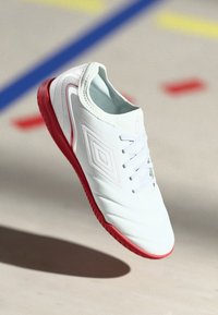 Weißer Sportschuh mit roter Sohle, ausgestattet mit einer strukturierten Oberseite, Schnürverschluss und einem rautenförmigen Logo an der Seite.