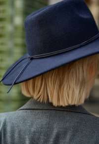 Cappello di feltro navy a tesa larga con corona piatta, dotato di una sottile banda abbinata e lacci. Superficie testurizzata, posizionato dietro il collo di una persona.