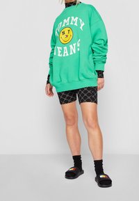 Felpa verde con vestibilità oversized, con il testo "TOMMY JEANS" e una grafica con un sorriso. Abbinata a pantaloni corti neri con motivo e sandali neri.