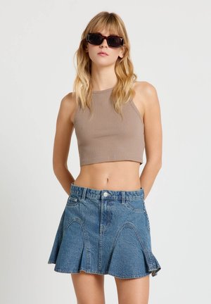 Donna che indossa un top beige senza maniche a vita corta, gonna di jeans svasata blu e occhiali da sole grandi e scuri, in piedi davanti a uno sfondo chiaro e semplice.