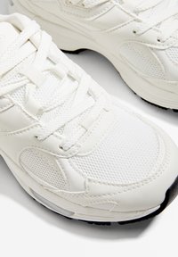Białe buty sportowe z mieszanki skórzanych i siateczkowych materiałów, o zaokrąglonym kształcie, z teksturowanymi akcentami oraz klasycznym sznurowanym zapięciem.