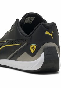 Musta ja harmaa urheilukenkä, jossa sileä pinta, keltaisia yksityiskohtia, rei'itetty muotoilu ja Ferrari-logo. Kenkässä on valkoinen pohja.