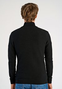 Svart strukturerad stickad tröja med hög krage och långa ärmar, med ribbade mudd och nederkant, sedd bakifrån.