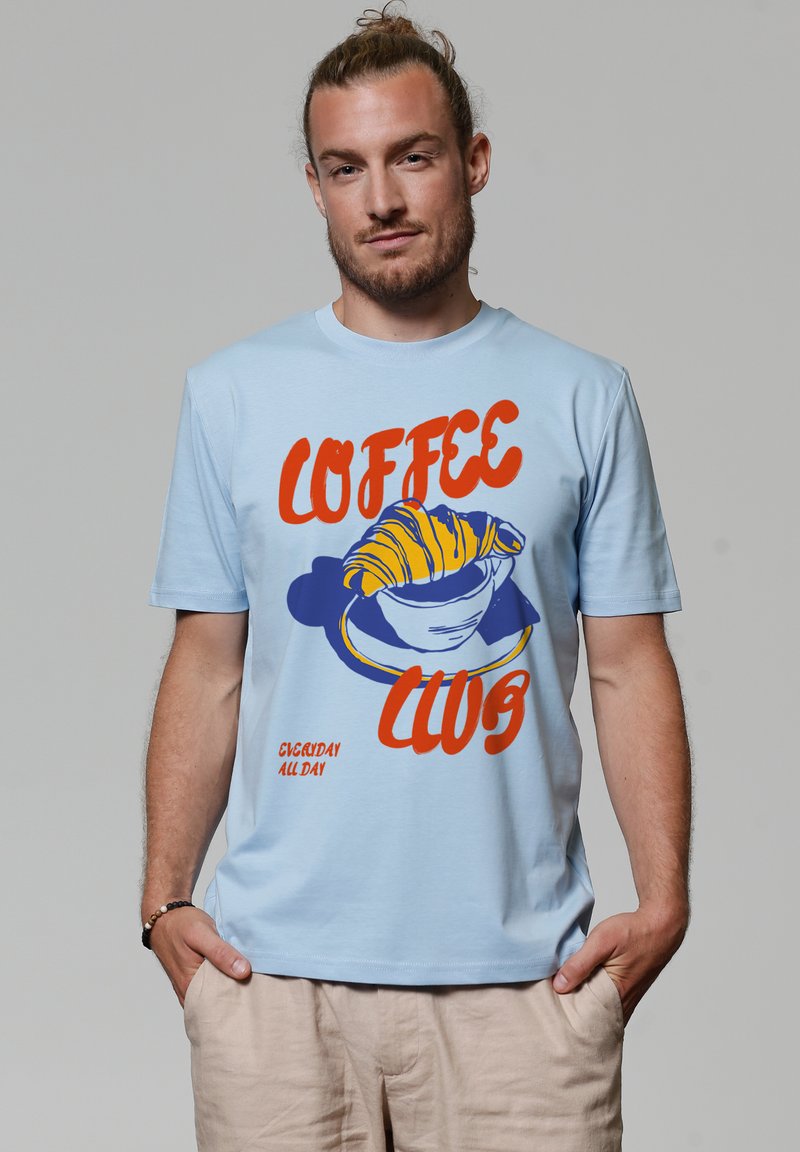 T-shirt à manches courtes bleu clair avec un design graphique d'une tasse de café et d'un croissant en orange et bleu. Texture de tissu douce.