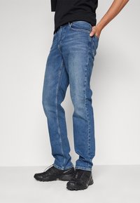 Jeans de mezclilla azul con un corte recto, que cuentan con una cremallera, bolsillos y un desgaste sutil en los muslos. Zapatillas negras completan el look.