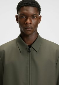 Olivgrön jacka med en slät textur, minimalistisk design och klassisk krage. Har en svart dragkedja framtill för stängning.