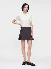 Femme aux cheveux courts et foncés portant une chemise blanche à manches courtes boutonnée, une jupe plissée foncée et des chaussures Mary Jane noires, debout avec la main sur la hanche.