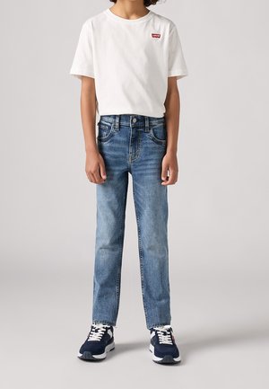 Barn iført hvid Levi's t-shirt, blå denimjeans og mørkeblå sneakers står foran en ensfarvet baggrund.