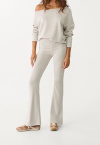 Femme portant un top beige à épaules dénudées et manches longues avec un pantalon évasé taille haute assorti et des baskets beiges avec des lacets blancs.