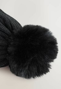 Bonnet en tricot noir avec un grand pompon en fausse fourrure noire sur le dessus. La texture est douce, avec un motif côtelé sur l'ensemble du corps.