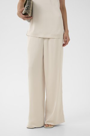 Kobieta nosząca beżowe spodnie o szerokich nogawkach i dopasowany bezrękawnik, trzymająca torebkę typu kopertówka w z wzorem skóry węża.