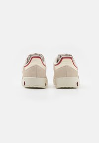 Beige suède sneakers met witte accenten, rode details en een getextureerde zool. De achteraanzicht toont een gewatteerde kraag en subtiele branding.