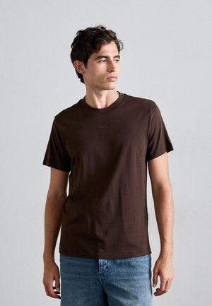 Mand iført en mørkebrun T-shirt med korte ærmer og "SANDRO" logo samt blå jeans, står op ad en ensfarvet lysegrå væg.