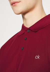 Červené polo tričko s textúrovanou tkaninou, tromi čiernymi gombíkmi a malým bielym výšivkou "ck" na ľavej hrudi.